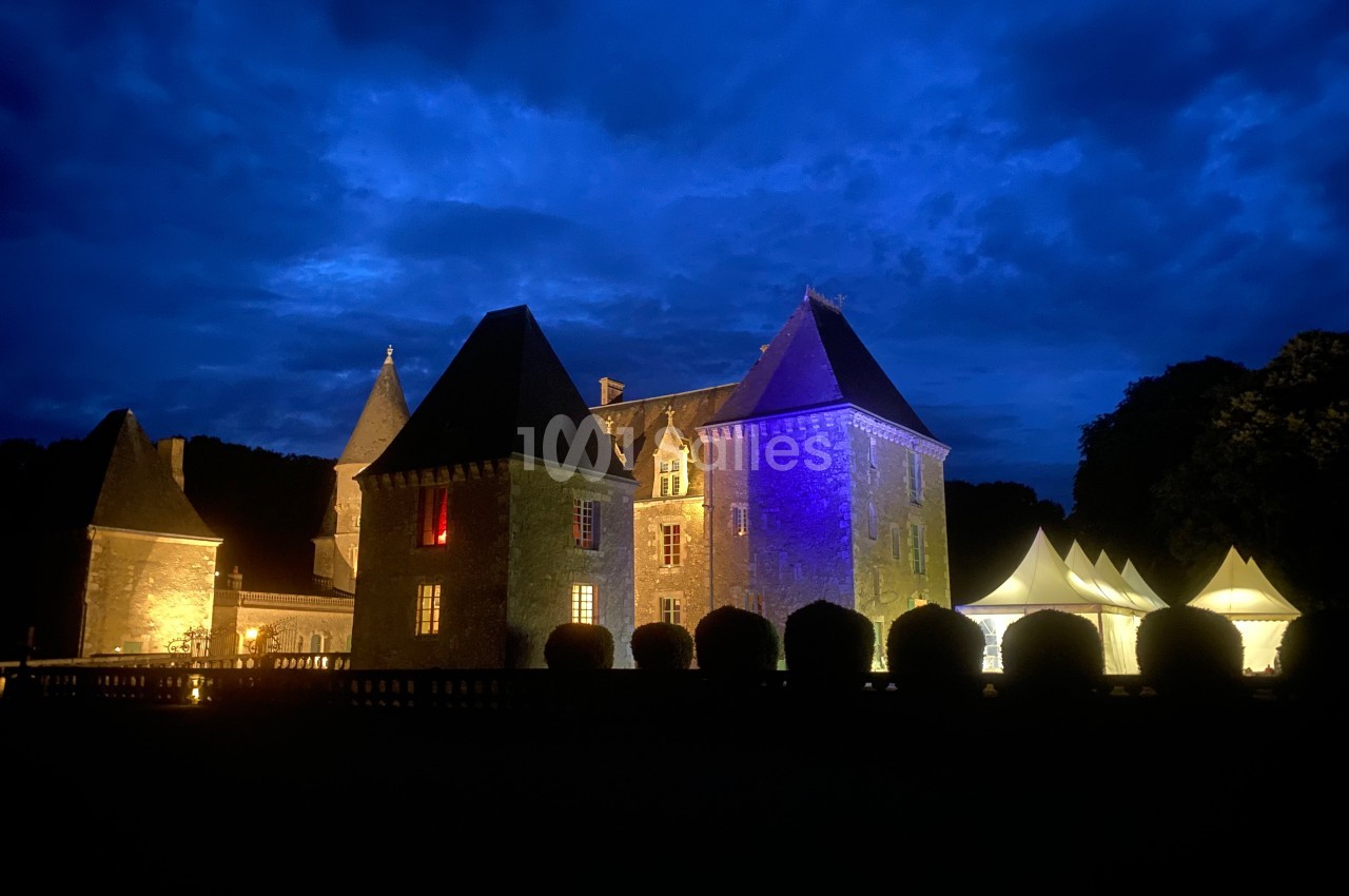 Château éclairé de nuit avec des lumières colorées, entouré de buissons et de tentes blanches sous un ciel nuageux.