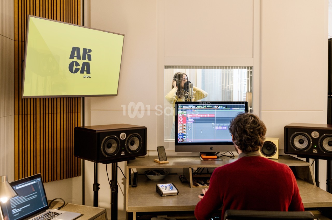 Un ingénieur du son travaille sur un projet audio devant un écran, tandis qu'une chanteuse enregistre derrière une vitre.