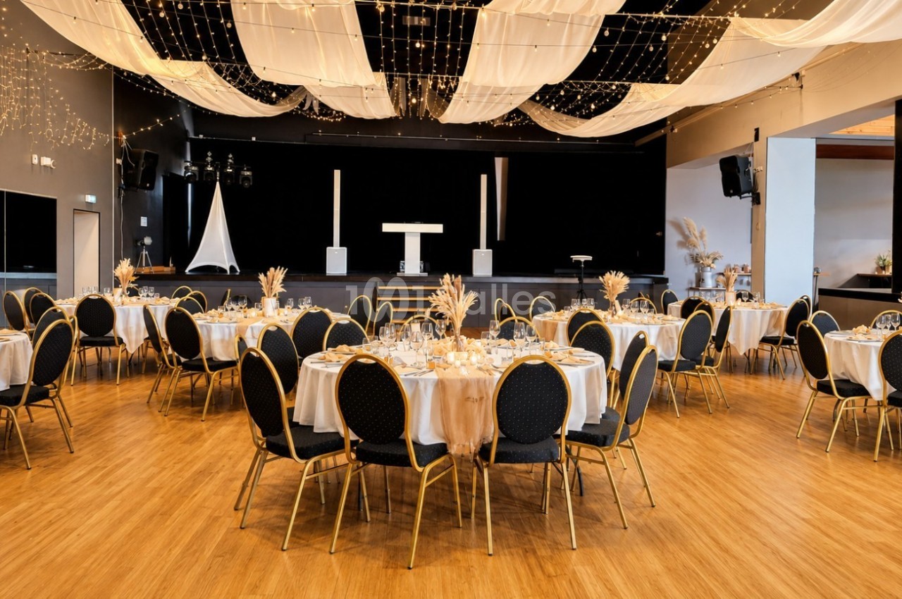 Salle de réception décorée avec des tables rondes dressées, des chaises noires et un plafond orné de guirlandes lumineuses.