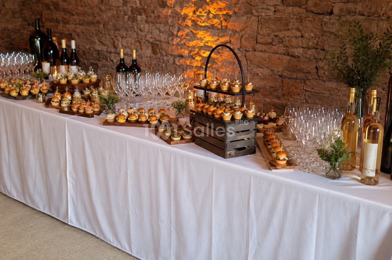 Table de buffet élégamment dressée avec verrines, bouteilles de vin et décorations végétales devant un mur en pierre.