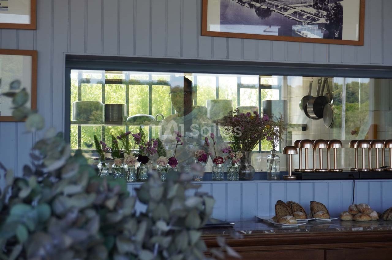 Un comptoir avec des fleurs en vase, des pâtisseries et un cuisinier visible à travers une fenêtre intérieure.
