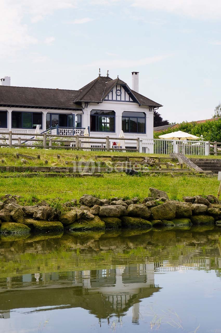 Maison à colombages avec terrasse, entourée de verdure, bordée par un plan d'eau et un muret de pierres.