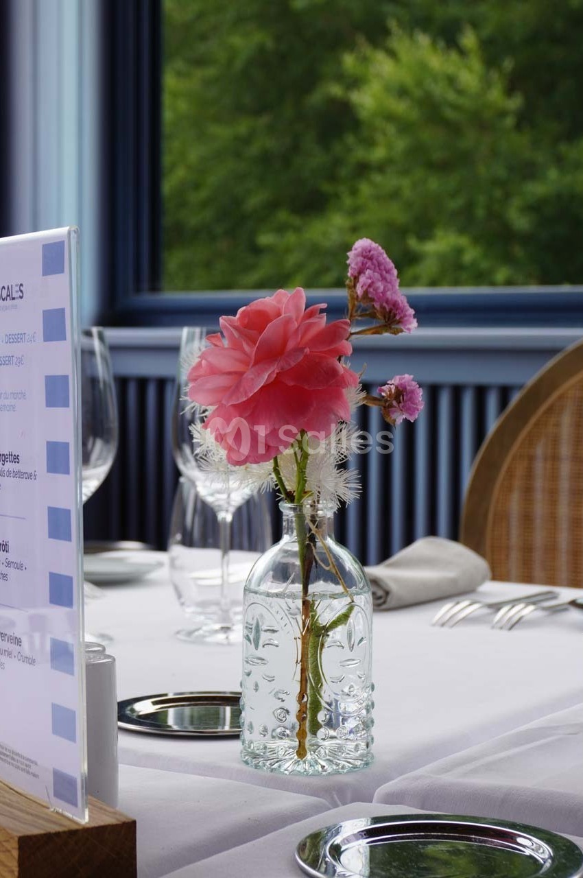 Un vase en verre contenant des fleurs roses et blanches posé sur une table dressée près d'une fenêtre donnant sur un jardin.