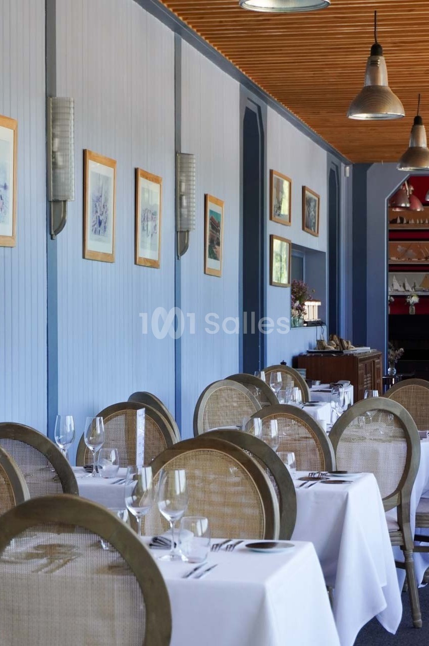 Salle de restaurant élégante avec tables dressées, nappes blanches, chaises en osier et décorations murales.
