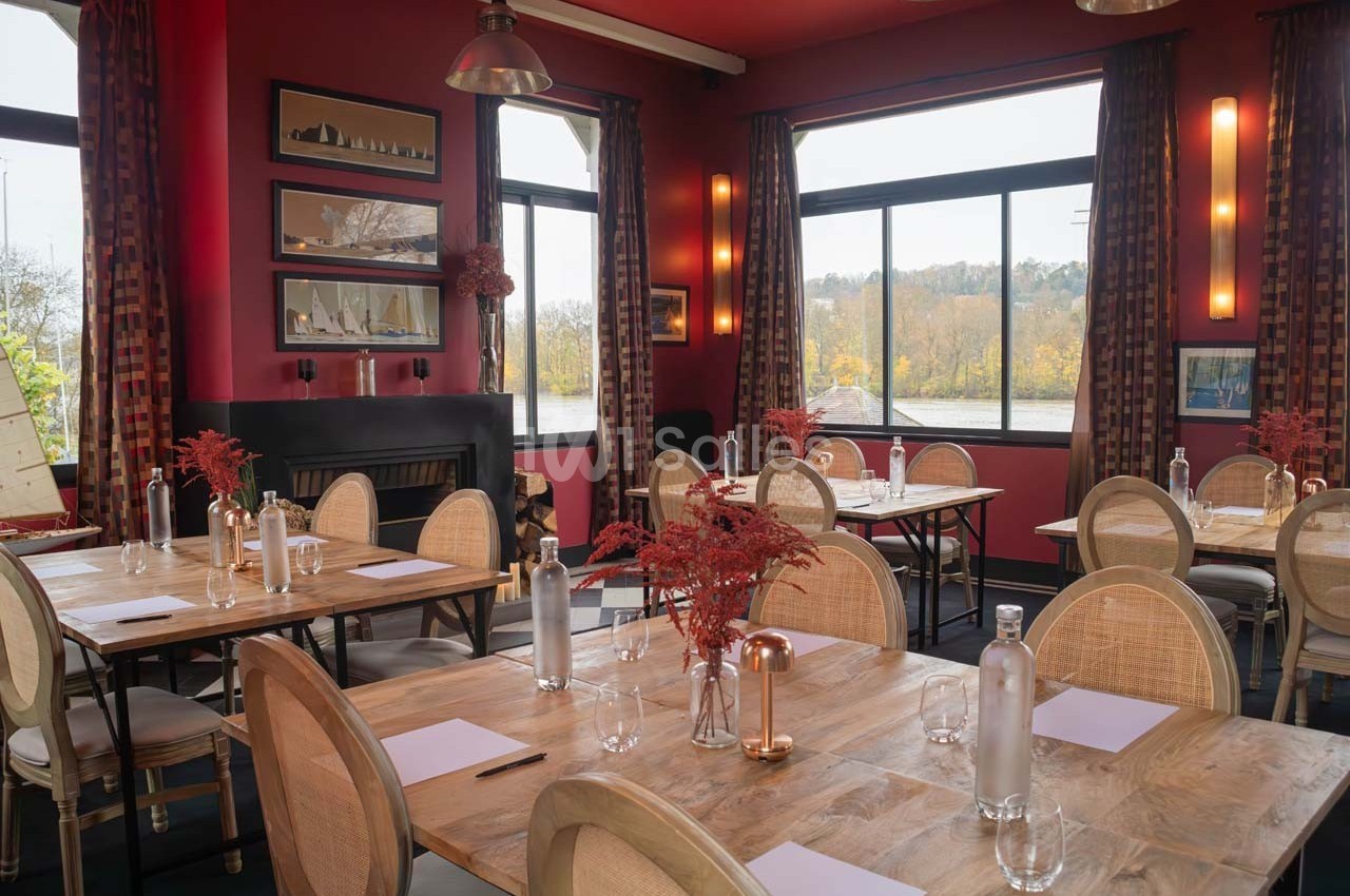 Salle de restaurant lumineuse avec tables en bois, chaises en rotin, décor rouge et grandes fenêtres donnant sur un paysage…