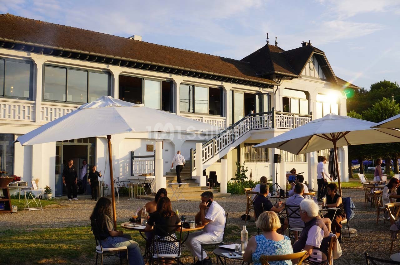 Personnes attablées en terrasse devant un grand bâtiment blanc avec baies vitrées, par une fin de journée ensoleillée.