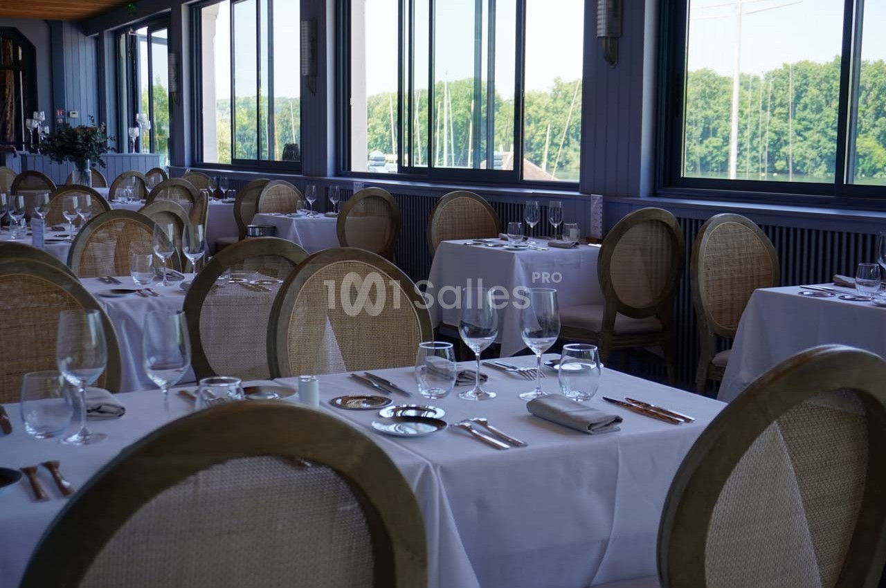 Salle de restaurant lumineuse avec tables dressées, nappes blanches et grandes fenêtres donnant sur un paysage verdoyant.