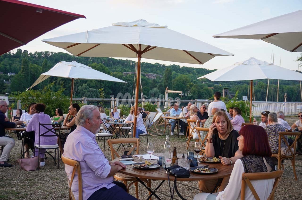 Personnes dînant en terrasse sous des parasols, avec vue sur un paysage verdoyant en arrière-plan.