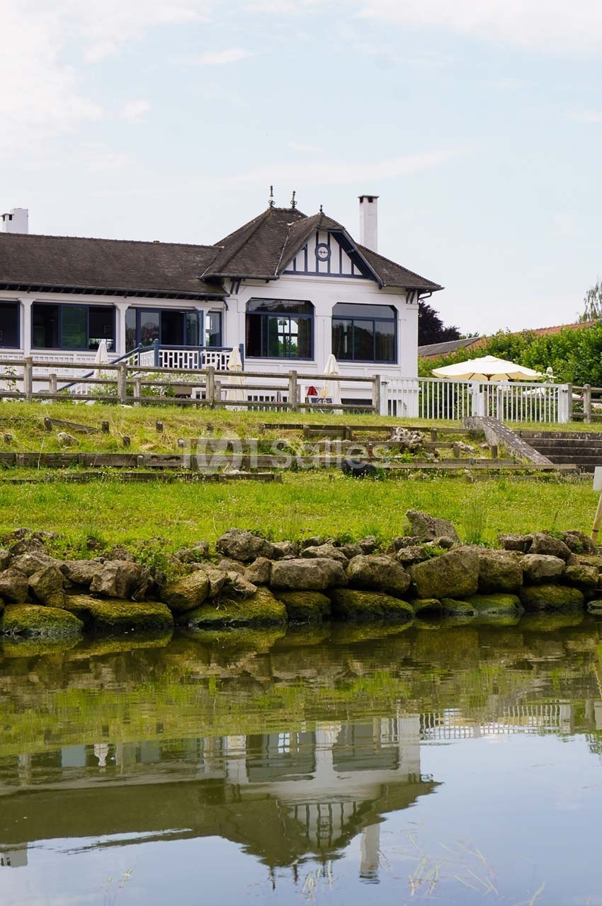 Maison à colombages avec terrasse, entourée de verdure, bordée par un plan d'eau et un muret de pierres.
