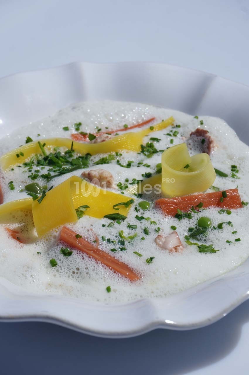 Assiette de soupe mousseuse garnie de légumes colorés, crevettes et herbes fraîches.