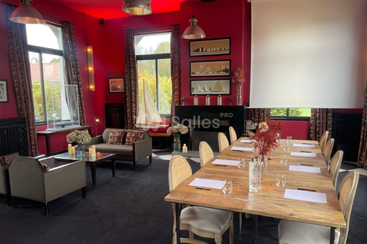 Salle de réunion lumineuse avec murs rouges, table en bois, chaises, coin salon et écran de projection.
