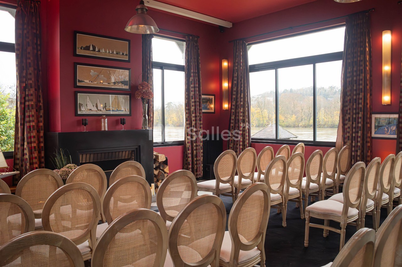 Salle de réunion lumineuse avec chaises alignées, murs rouges, grandes fenêtres donnant sur un paysage arboré.