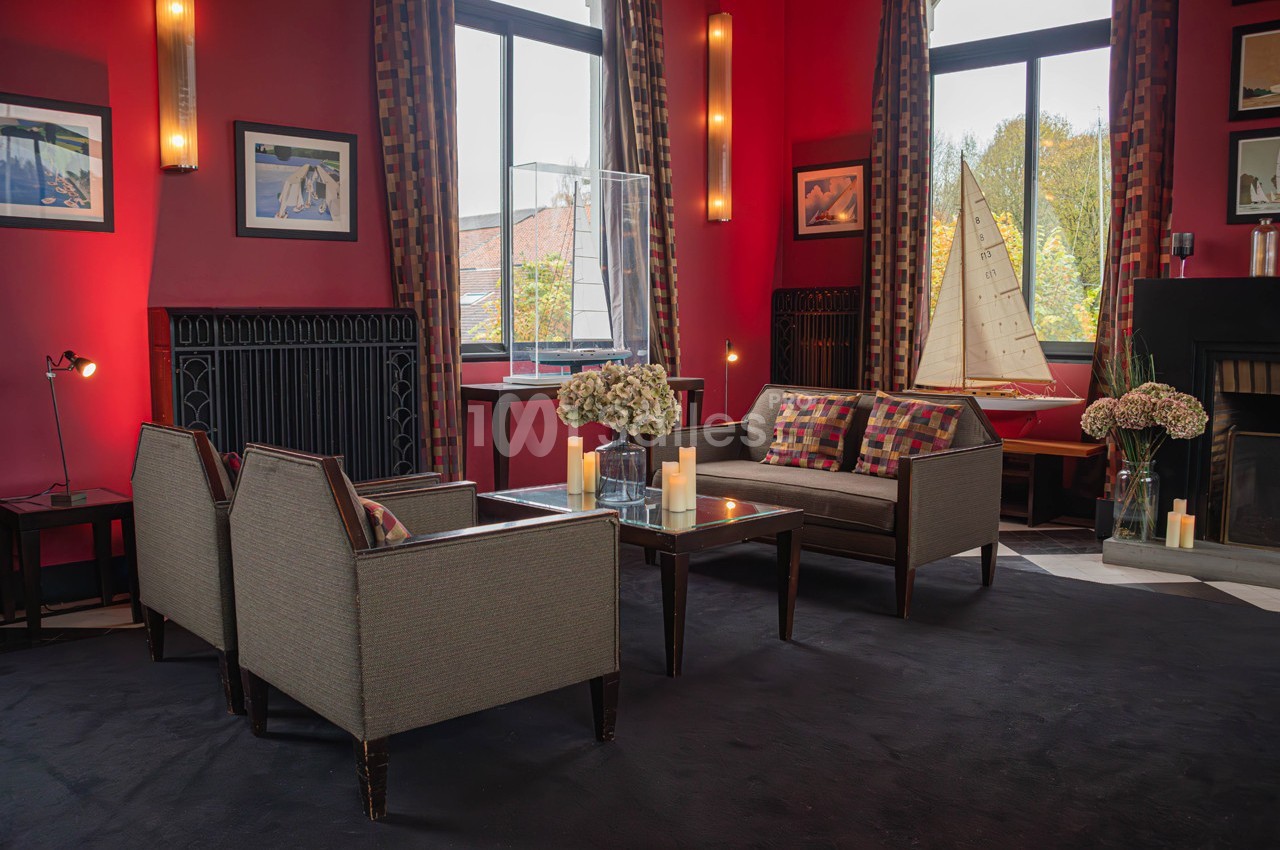 Salon chaleureux avec fauteuils, table basse, bougies, grandes fenêtres, rideaux colorés et décoration nautique.