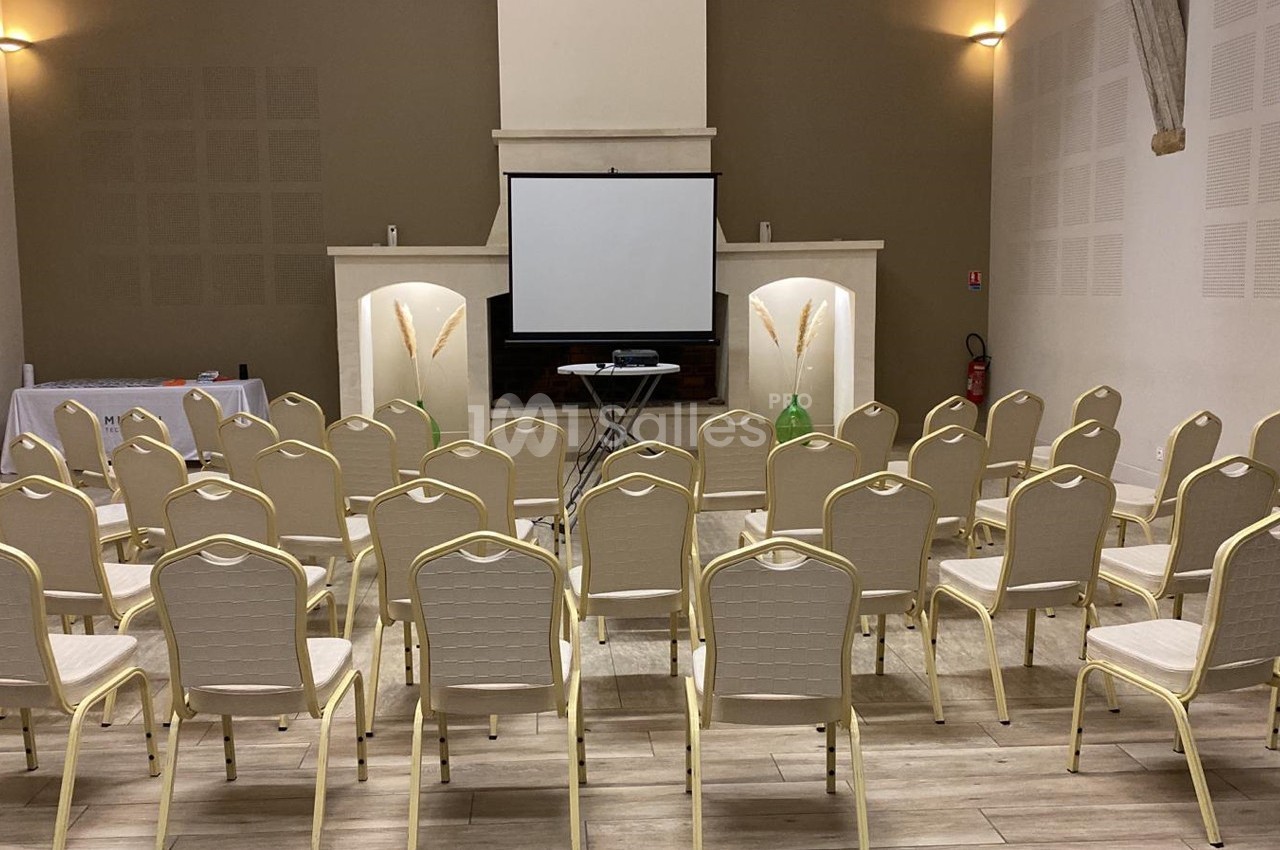 Salle de conférence avec des rangées de chaises beige alignées face à un écran de projection et un pupitre.