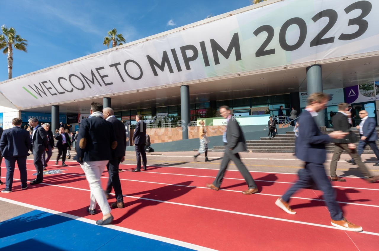 Des personnes marchent devant un bâtiment avec une grande bannière indiquant ’Welcome to MIPIM 2023’.