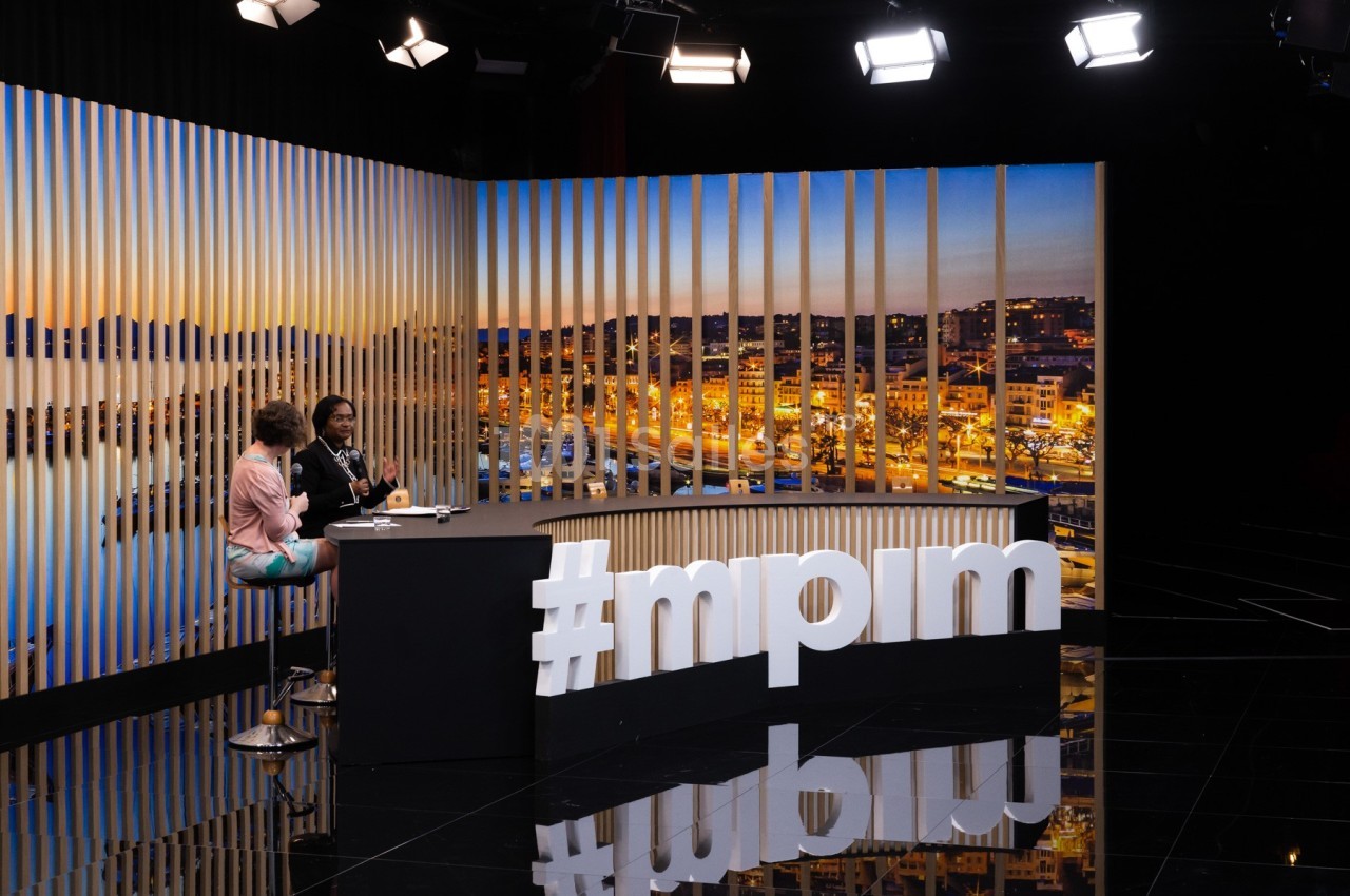 Deux personnes discutent sur un plateau télévisé avec vue nocturne sur une ville en arrière-plan.