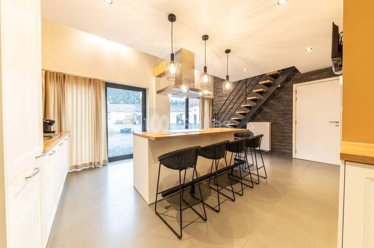 Cuisine moderne avec îlot central, tabourets noirs, luminaires suspendus et escalier en bois menant à l'étage.