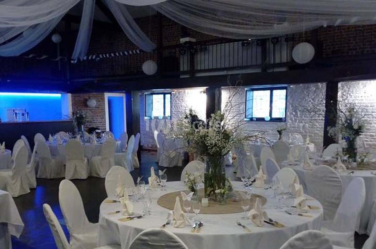 Salle de réception décorée avec des tables rondes, nappes blanches, chaises houssées et arrangements floraux.