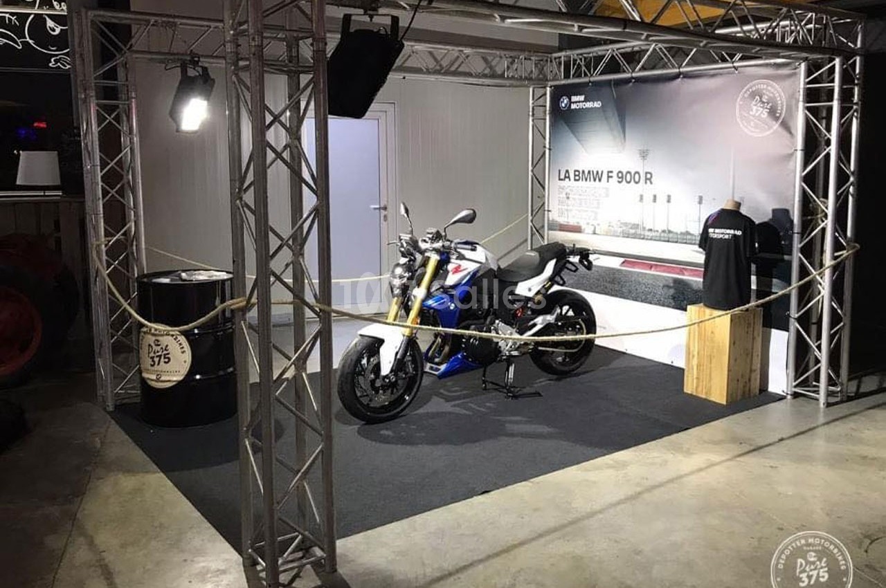 Stand d'exposition avec une moto BMW F 900 R, éclairage suspendu et décor industriel en arrière-plan.