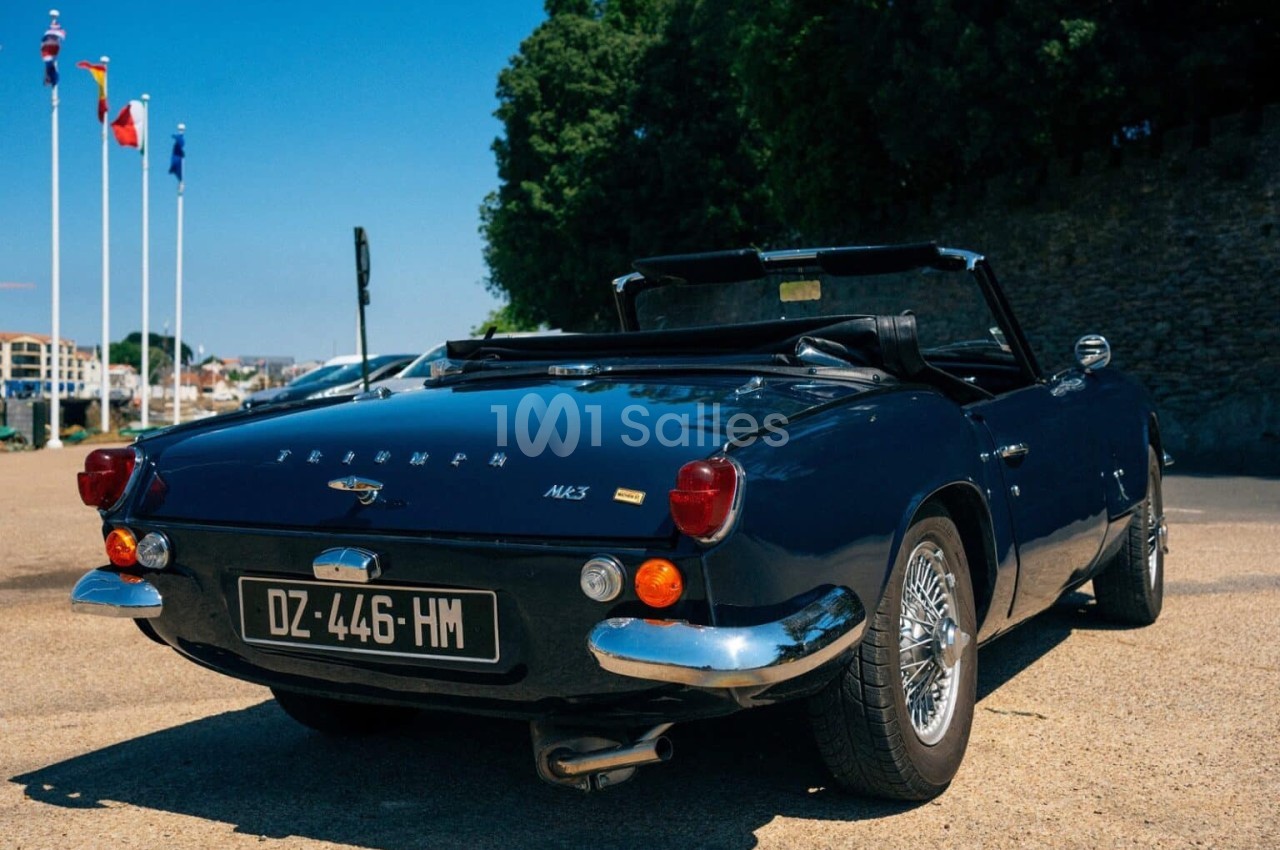 Voiture décapotable classique bleue garée sur une esplanade, avec des drapeaux et des arbres en arrière-plan.