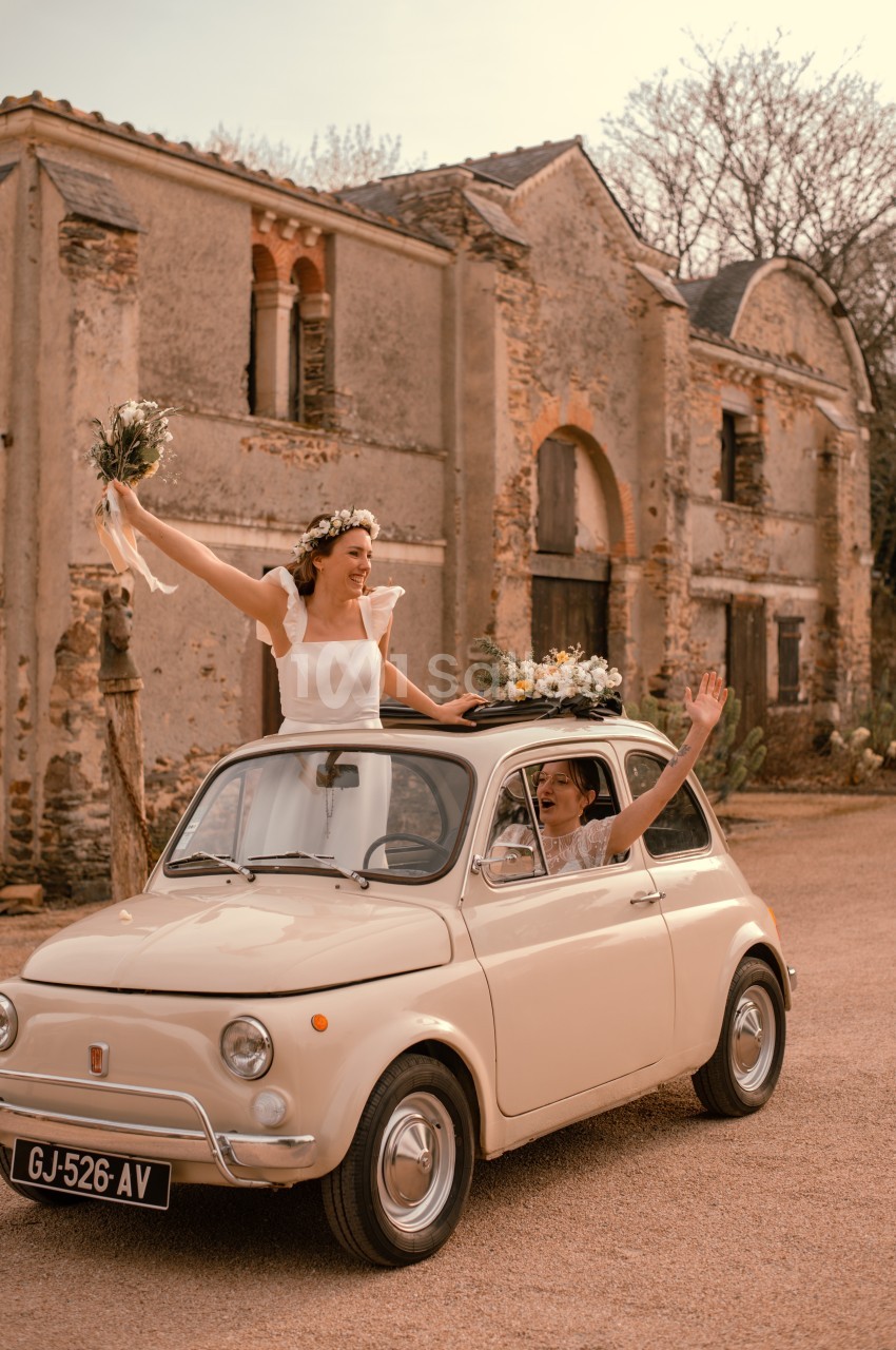Une mariée souriante agite un bouquet depuis une petite voiture ancienne devant un bâtiment en pierre.