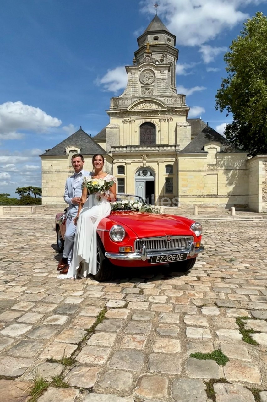Un couple en tenue de mariage pose devant une voiture rouge ancienne, avec une église en arrière-plan.
