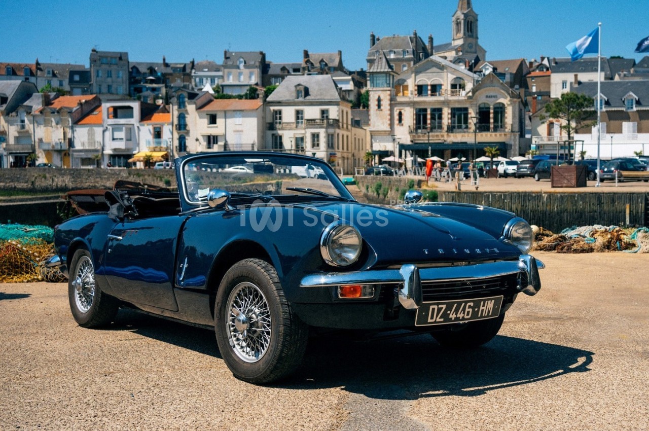 Voiture décapotable classique noire stationnée sur un quai, avec des bâtiments et une église en arrière-plan.