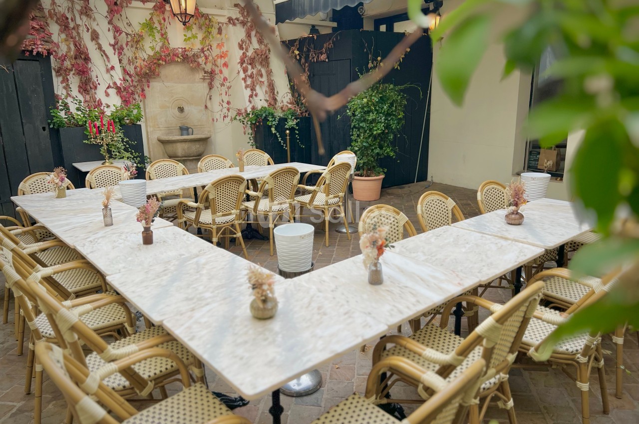 Terrasse extérieure avec tables en marbre disposées en U, chaises en rotin, plantes et fontaine murale en arrière-plan.