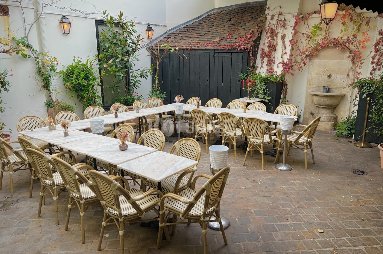 Terrasse pavée avec tables en marbre, chaises en rotin, plantes grimpantes et une fontaine murale en arrière-plan.