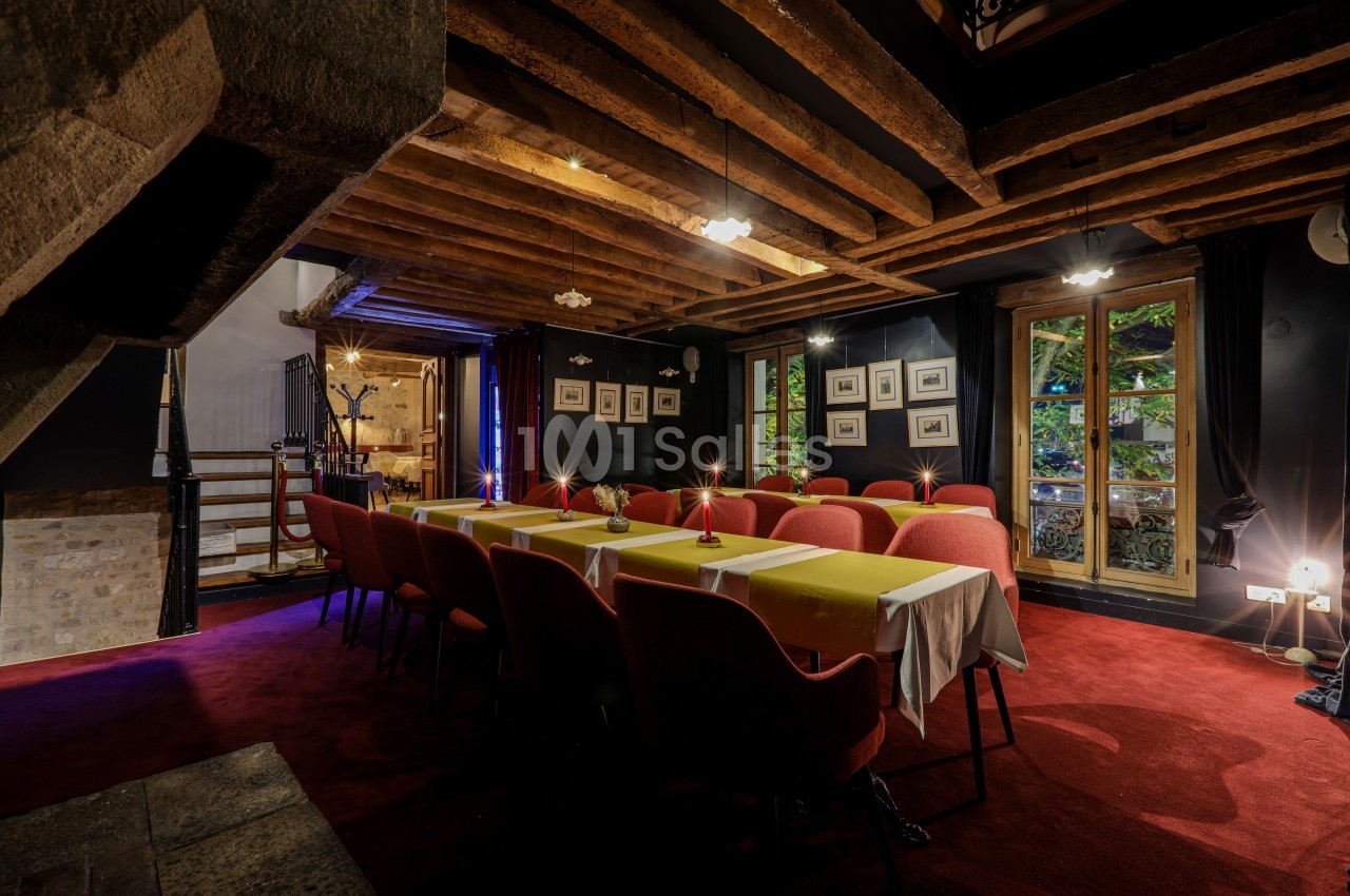 Salle de réunion chaleureuse avec poutres apparentes, tables dressées, chaises rouges et fenêtres donnant sur un jardin.