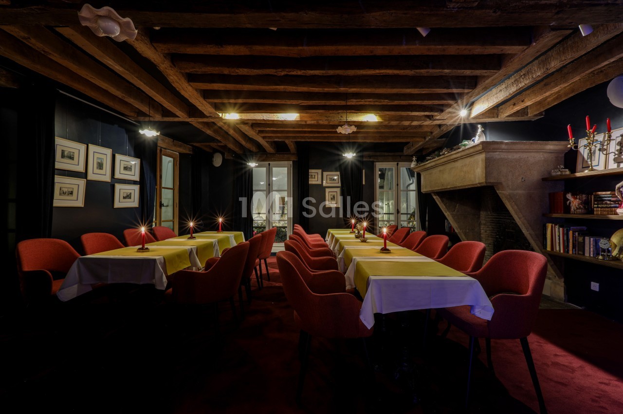 Salle à manger chaleureuse avec poutres apparentes, tables dressées, chaises rouges et cheminée en pierre.