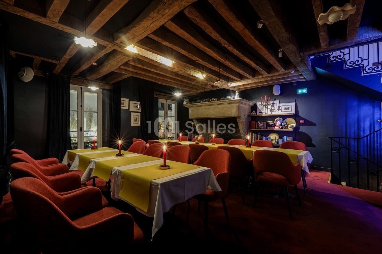 Salle à manger chaleureuse avec poutres apparentes, tables dressées de nappes jaunes et cheminée en pierre.