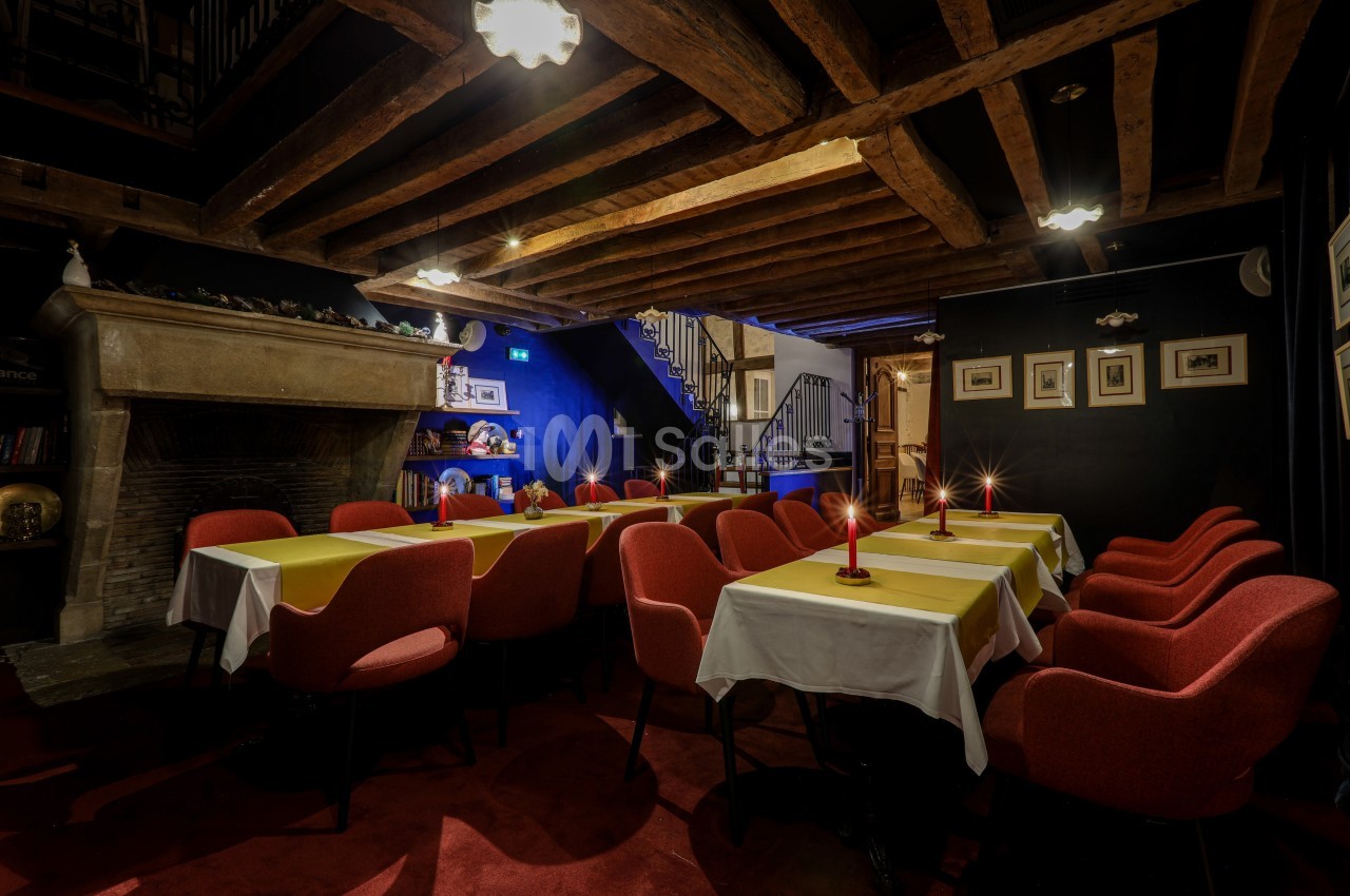 Salle de restaurant chaleureuse avec tables dressées, chaises rouges, poutres apparentes et éclairage tamisé.