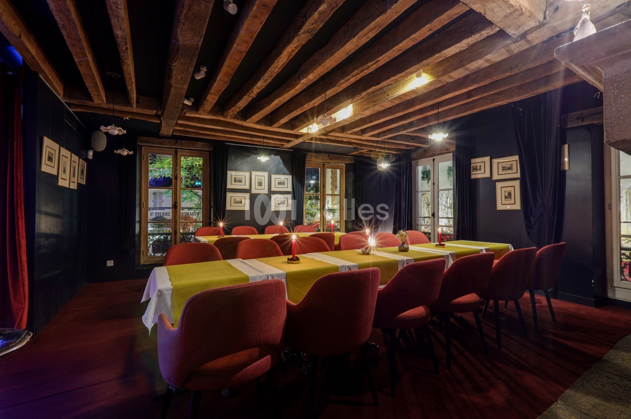 Salle de réunion chaleureuse avec poutres apparentes, tables dressées et chaises rouges, éclairée par des fenêtres.