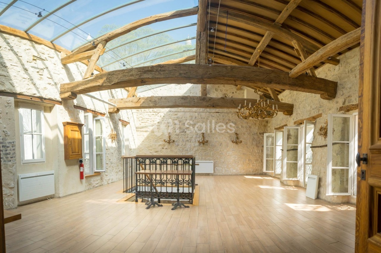 Salle lumineuse avec poutres apparentes, parquet en bois, murs en pierre et plafond en verre laissant entrer la lumière…