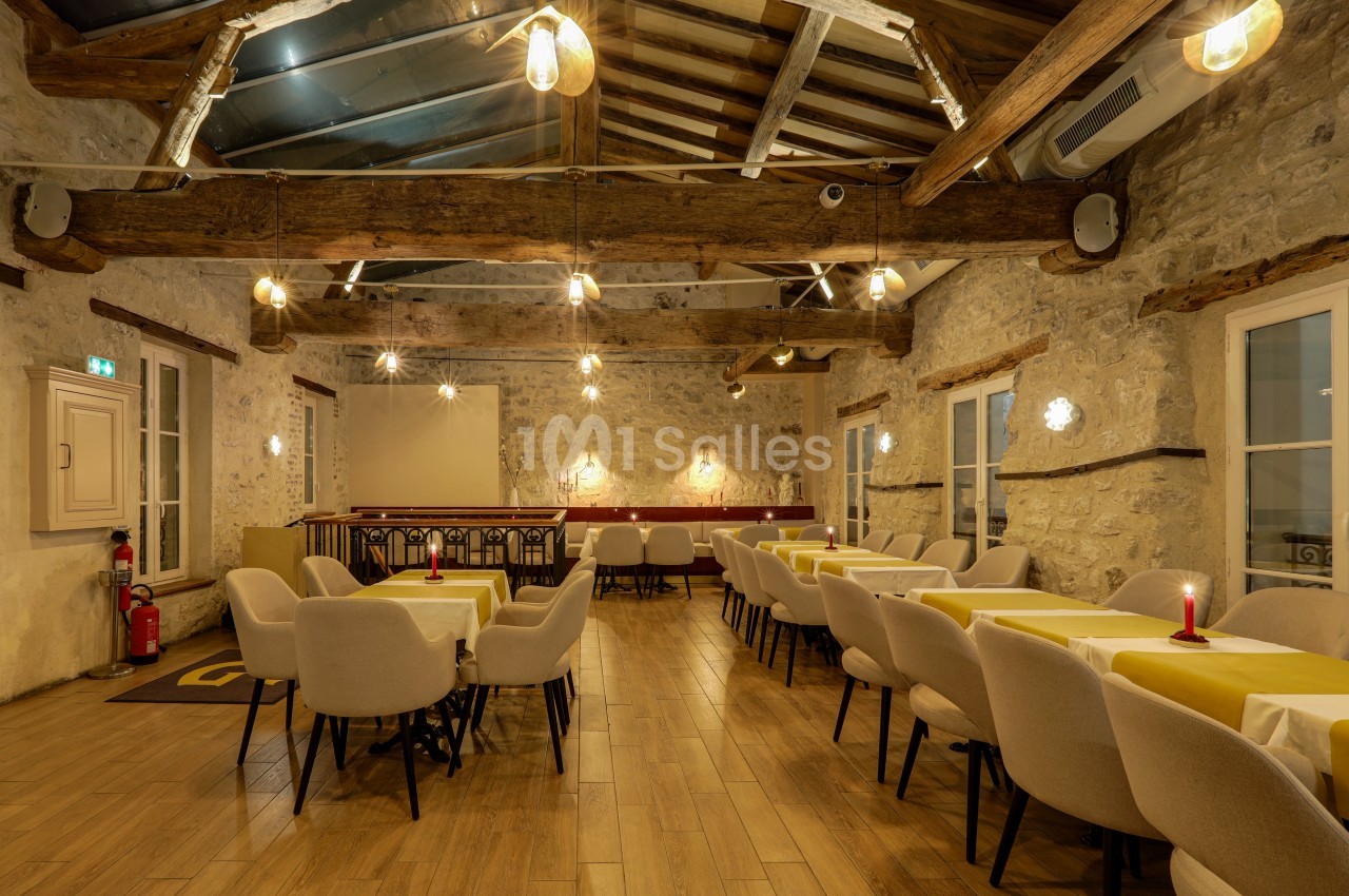 Salle de restaurant chaleureuse avec murs en pierre, poutres apparentes, tables dressées et éclairage tamisé.