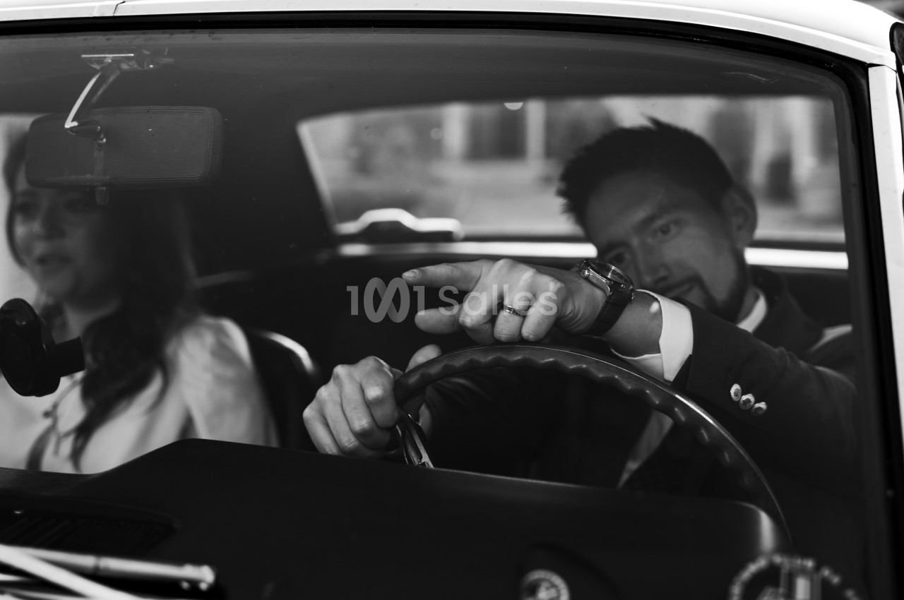 Un homme en costume conduit une voiture ancienne, accompagné d'une femme assise sur le siège passager.