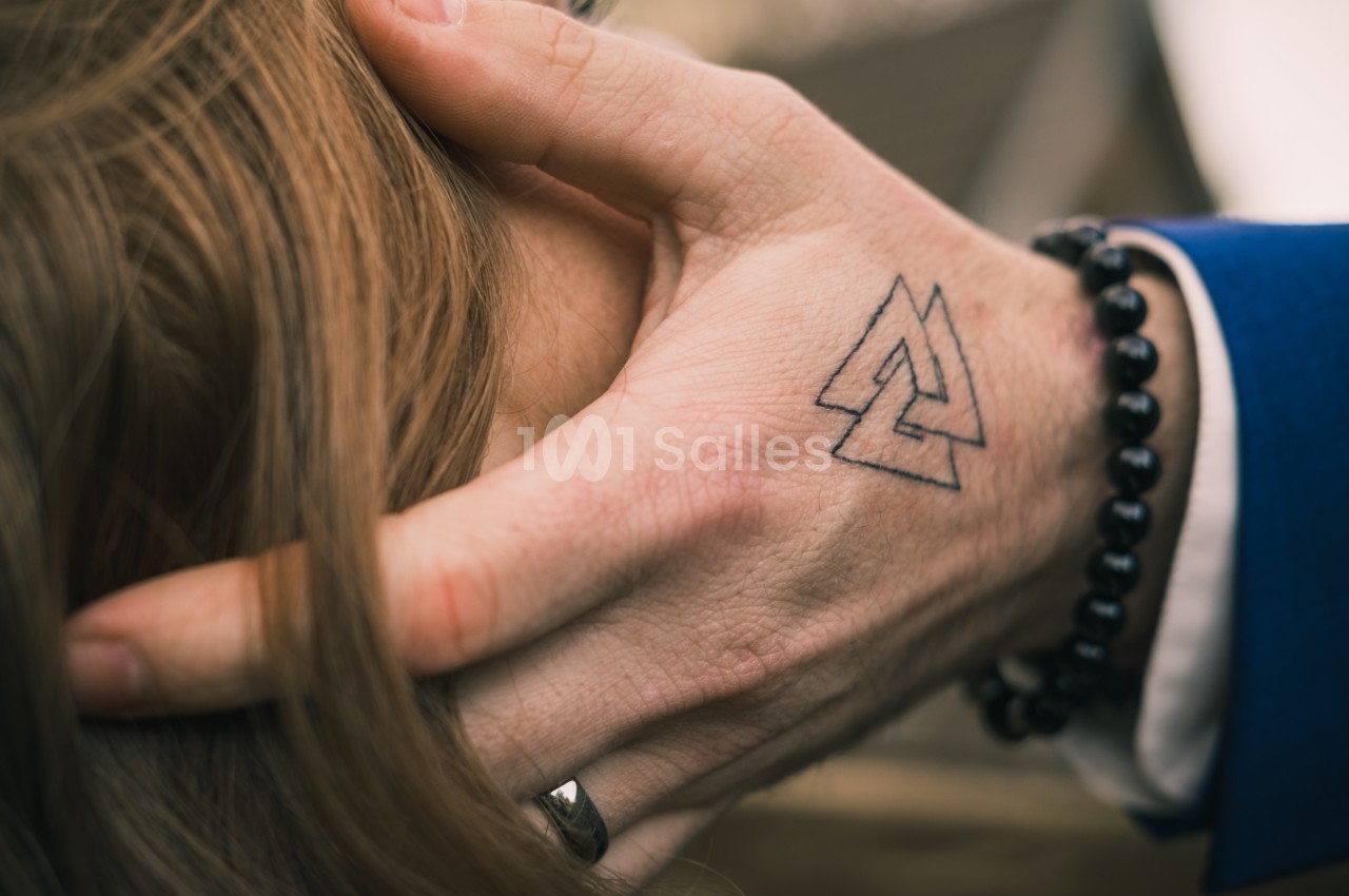 Main avec un tatouage de triangles entrelacés, posée sur des cheveux bruns, bracelet en perles noires visible.