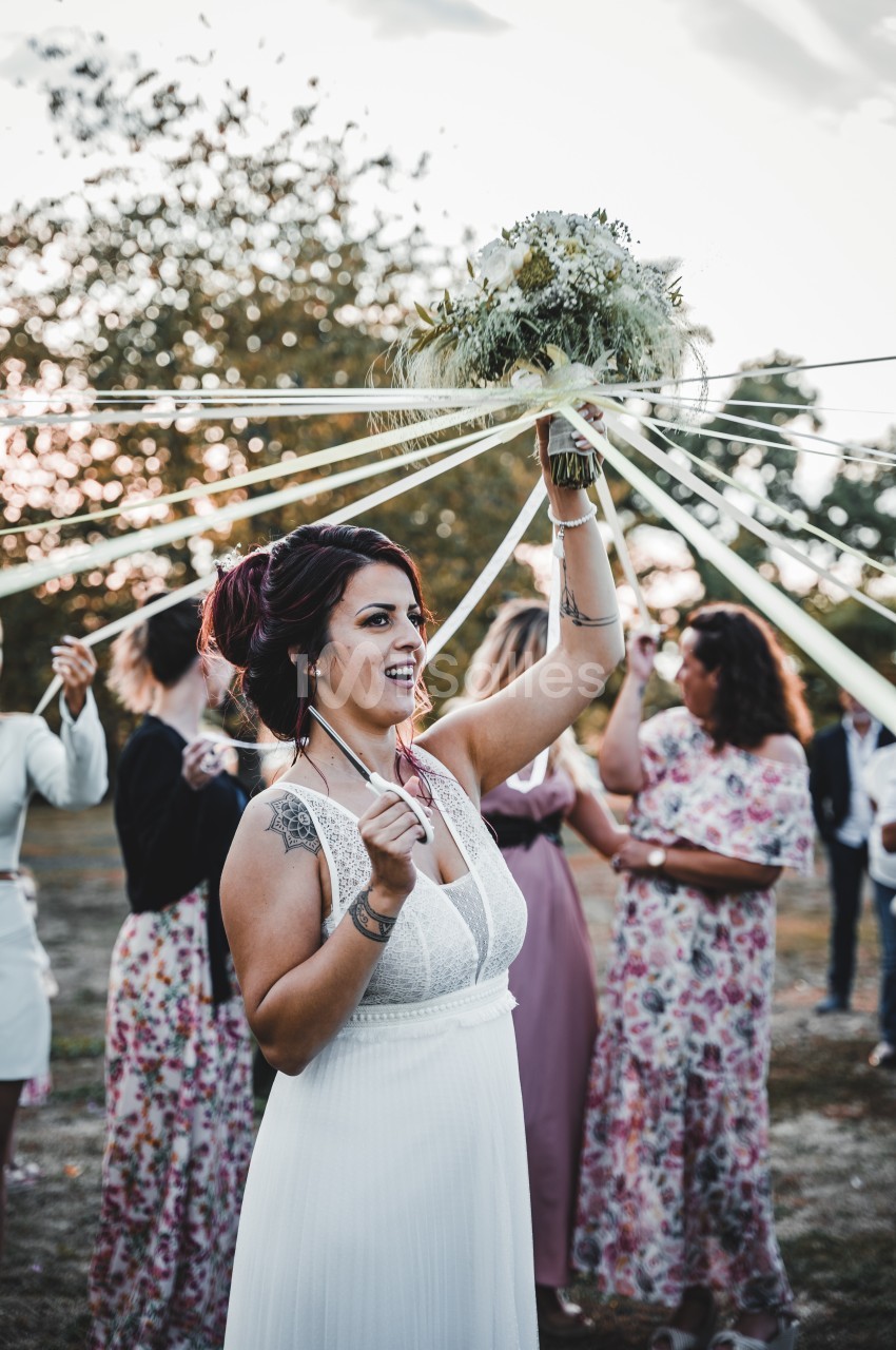 Une femme en robe blanche tient un bouquet entouré de rubans, entourée de personnes lors d'une cérémonie en extérieur.