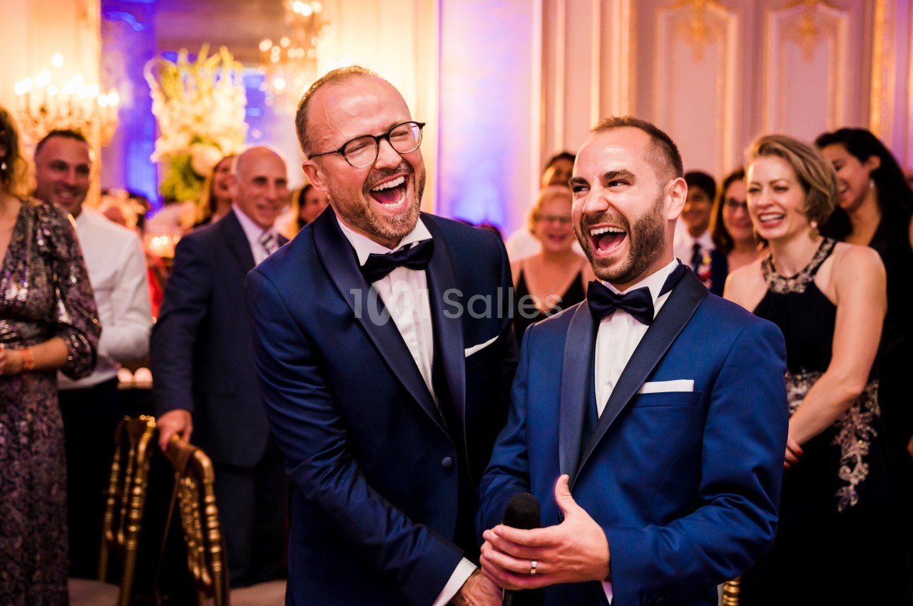 Deux hommes en costume souriant et riant lors d'un événement festif entourés d'invités élégants.
