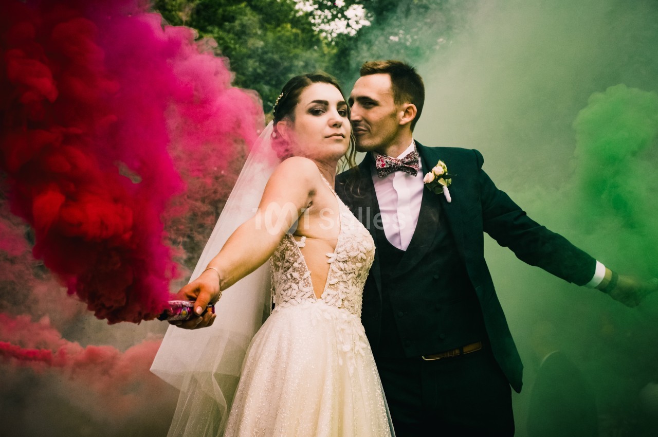 Un couple en tenue de mariage pose avec des fumigènes colorés rouge et vert dans un décor extérieur.