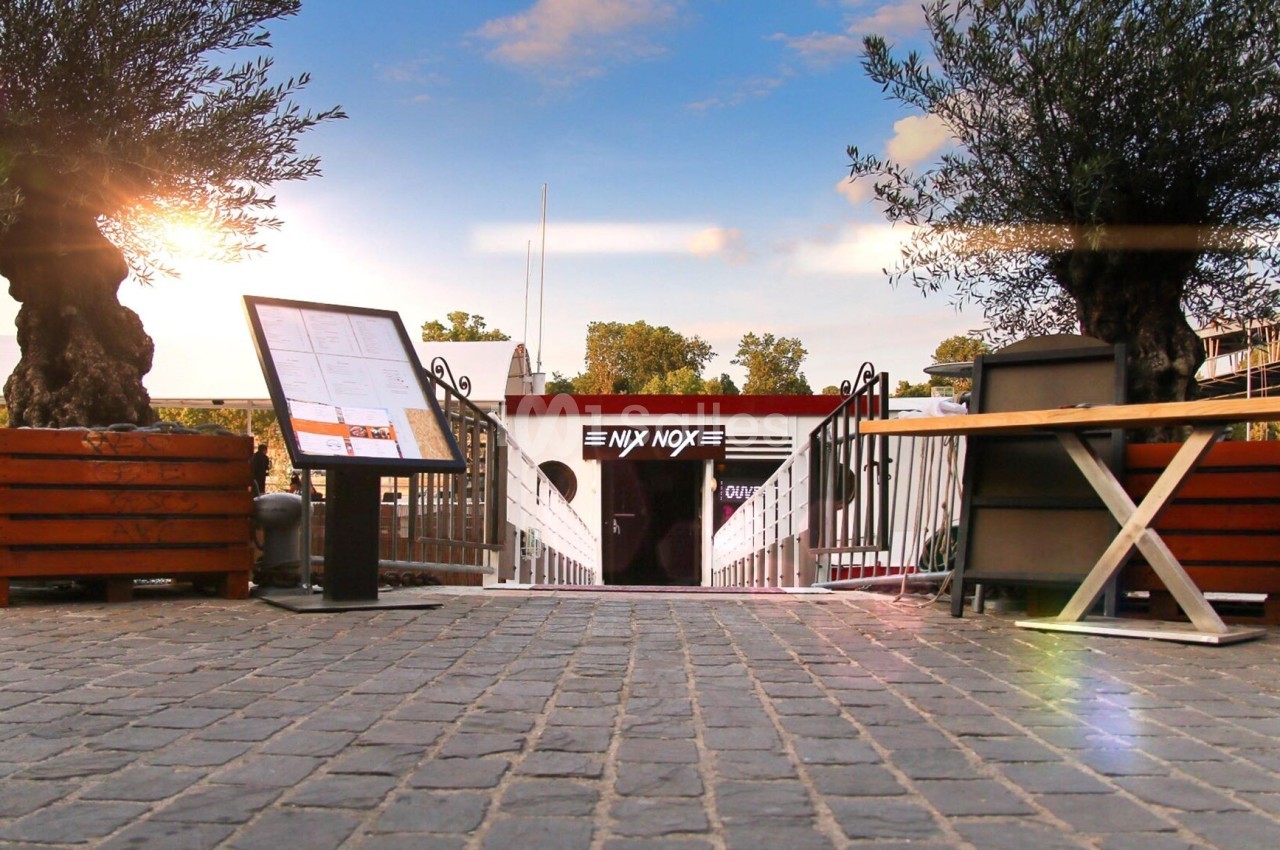 Terrasse extérieure avec oliviers, bancs en bois, tables et entrée de restaurant au coucher de soleil.