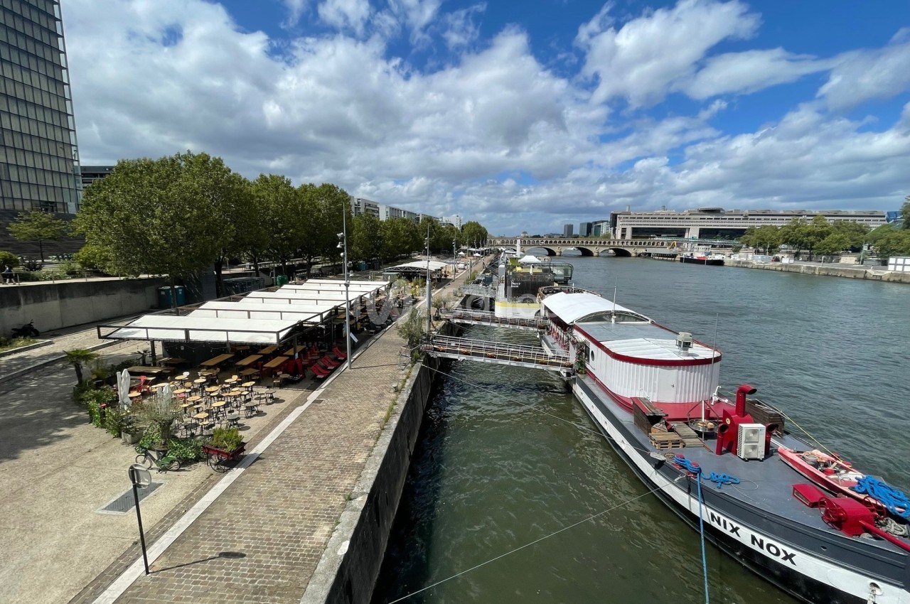 Terrasse en bord de Seine avec tables sous voiles blanches, chaises jaunes et bateau rouge amarré au quai.