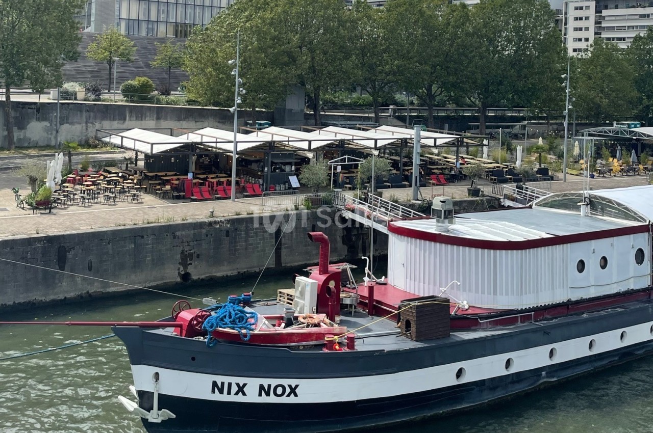 Bateau aménagé sur le quai avec terrasse couverte rouge et blanc, bâtiments en arrière-plan.