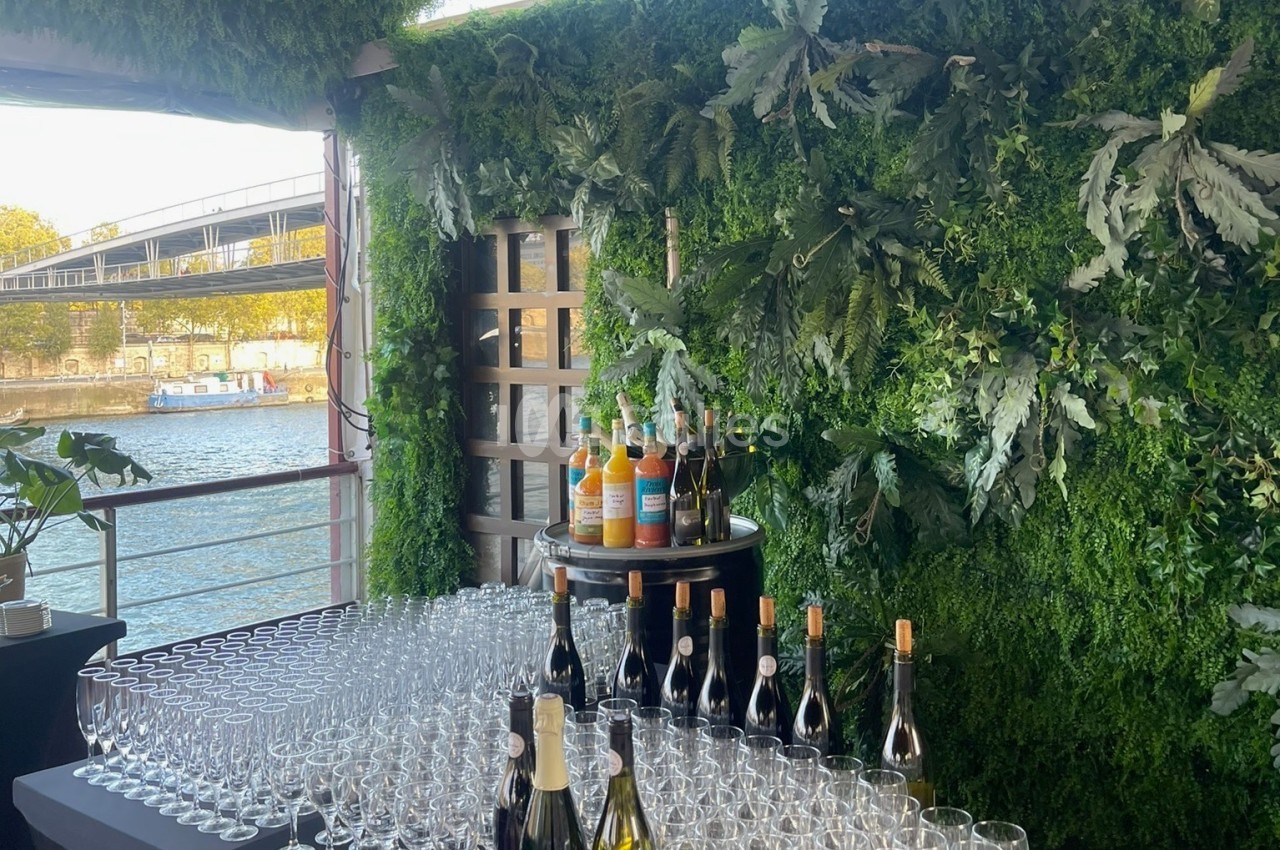 Espace de réception flottant en bord de Seine avec verres alignés, bouteilles de vin et jus, murs de verdure.