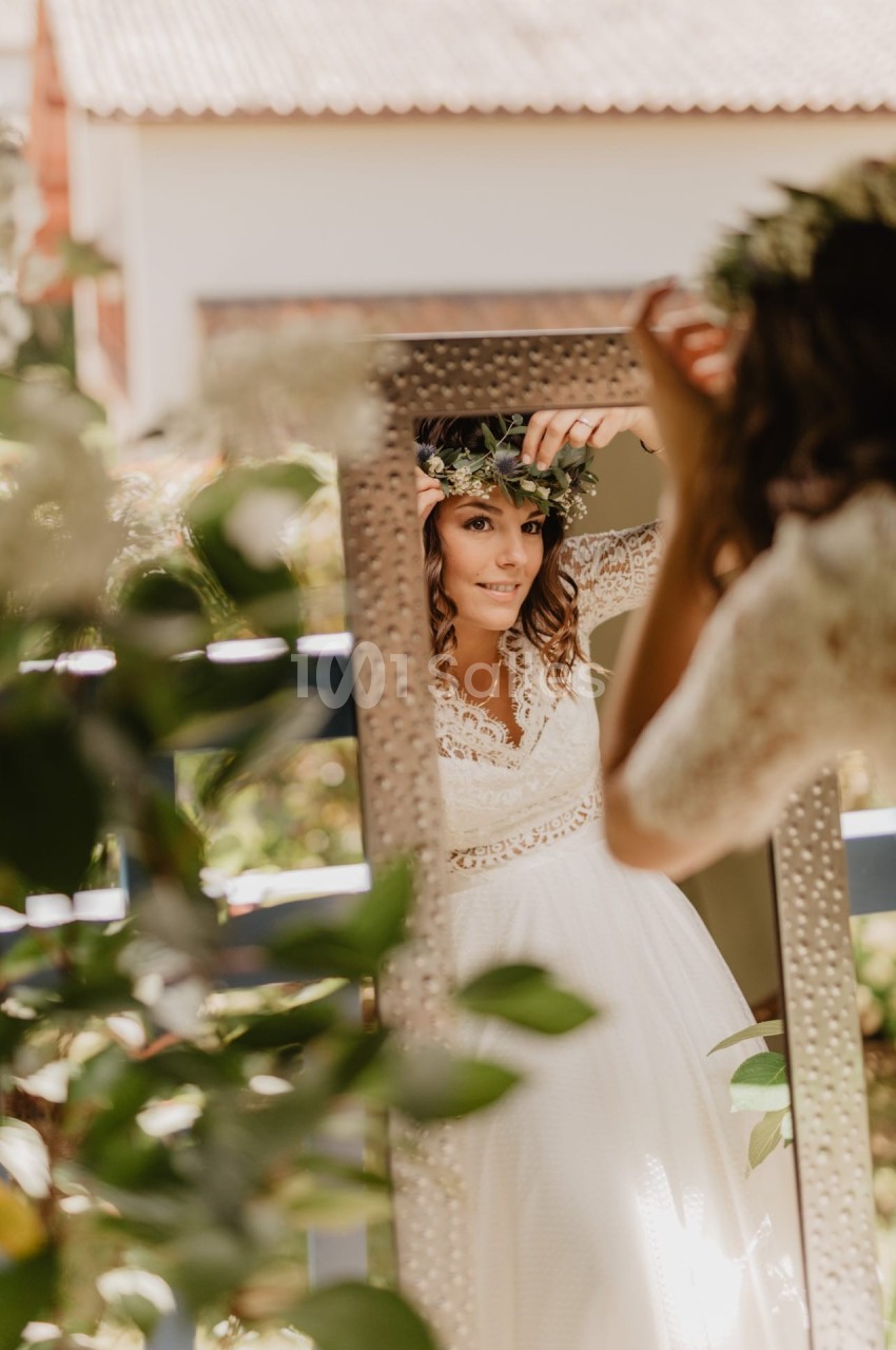 Une femme en robe de mariée ajuste une couronne de fleurs devant un miroir, entourée de verdure.