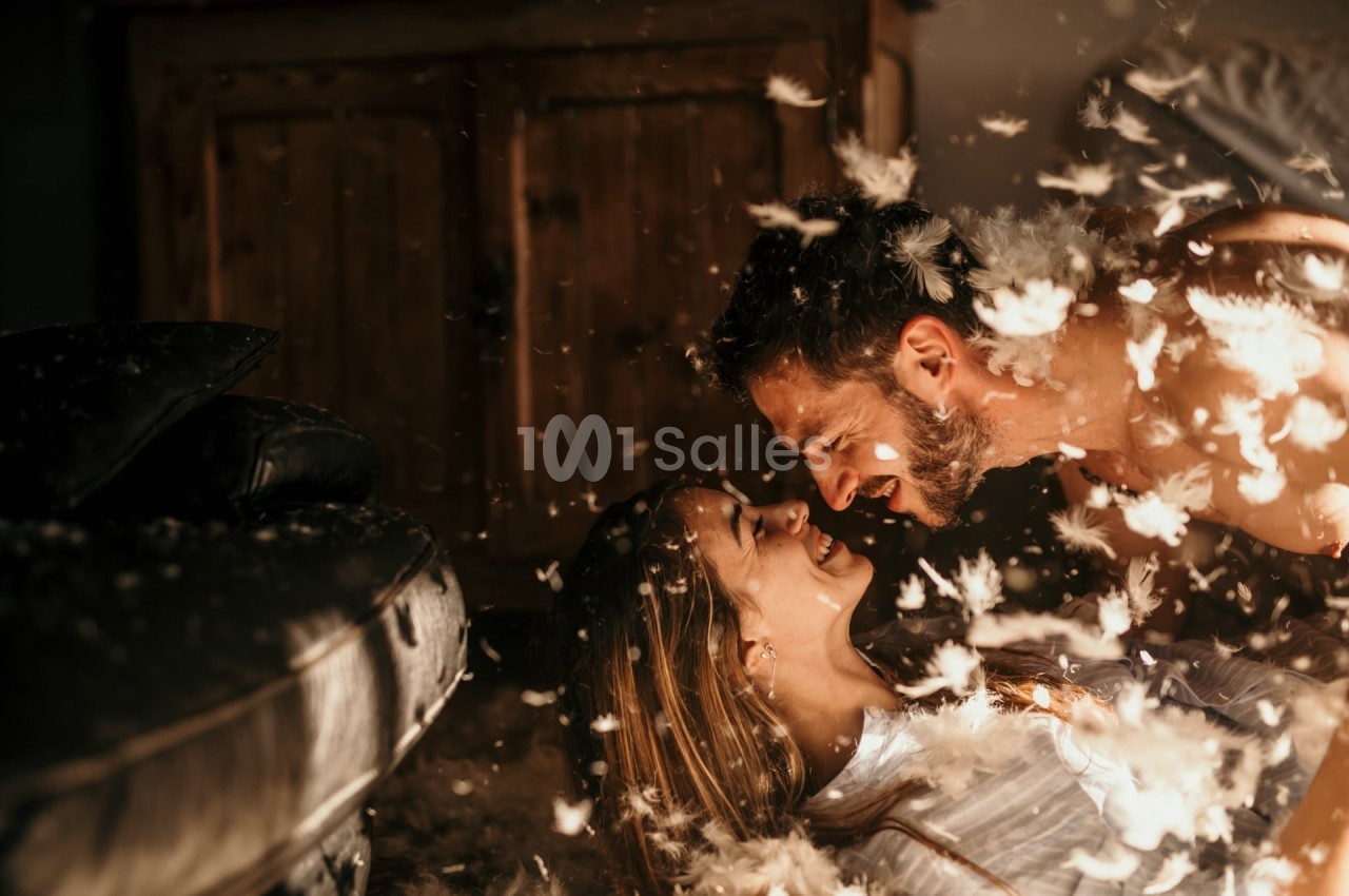 Un couple souriant allongé au sol entouré de plumes dans une ambiance chaleureuse et lumineuse.