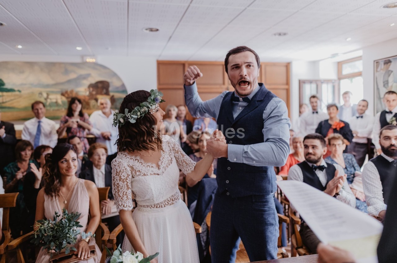 Un couple célèbre son mariage dans une salle avec des invités applaudissant en arrière-plan.