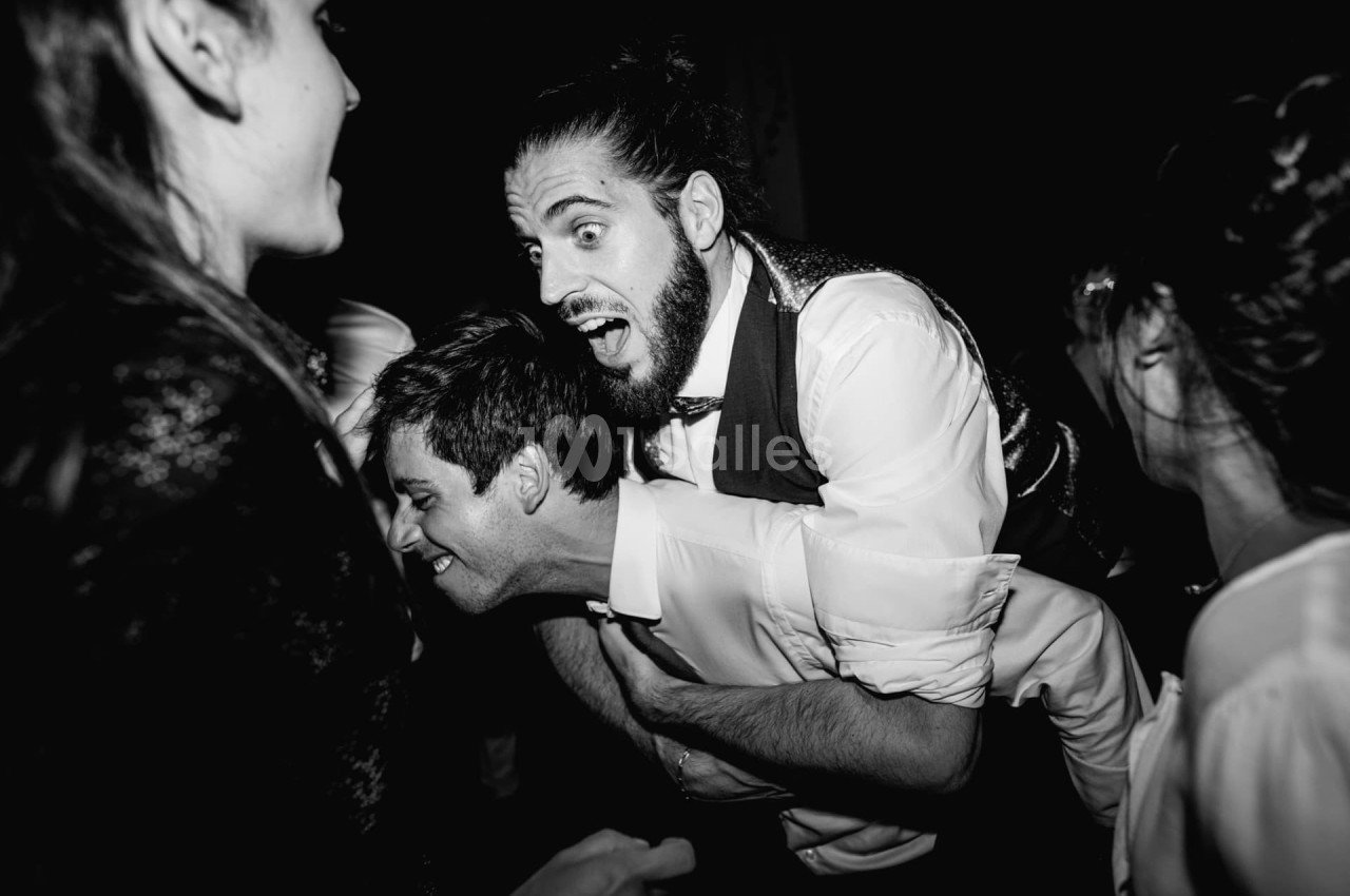 Deux hommes rient et jouent lors d'une fête, entourés d'autres personnes dans une ambiance festive.