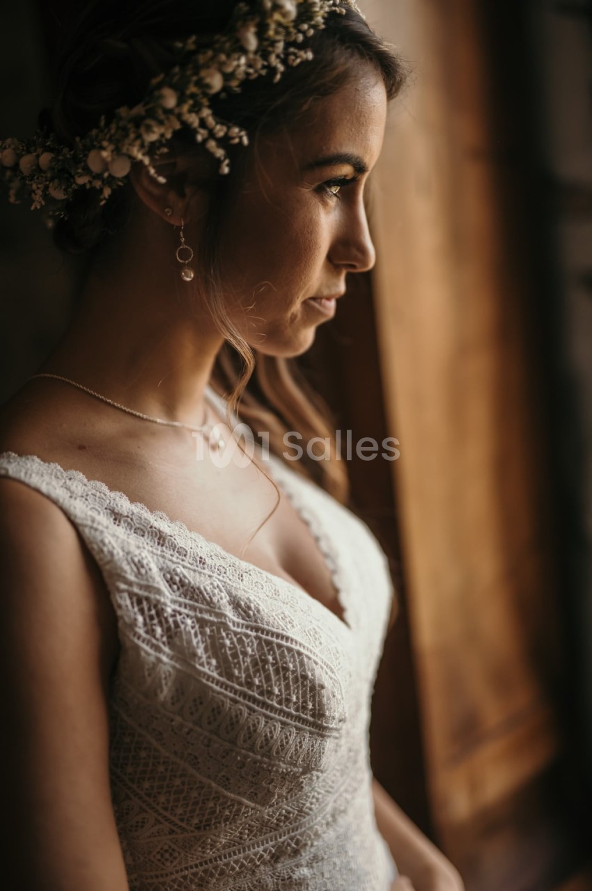 Femme en robe de mariée en dentelle blanche, coiffée d'une couronne florale, regardant par une fenêtre en bois.